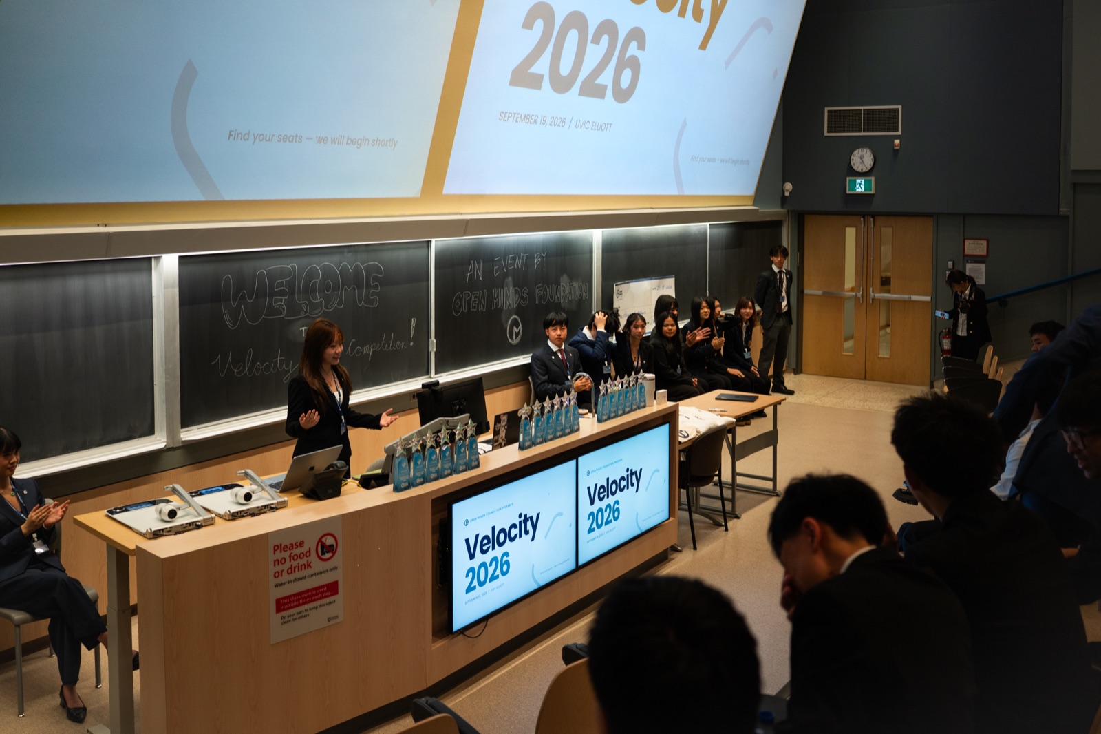 Opening remarks at Velocity 2026 in a University of Victoria lecture hall, with the organizing team at the front, award trophies on the desk, and “Welcome to Velocity Case Competition” written on the chalkboard
